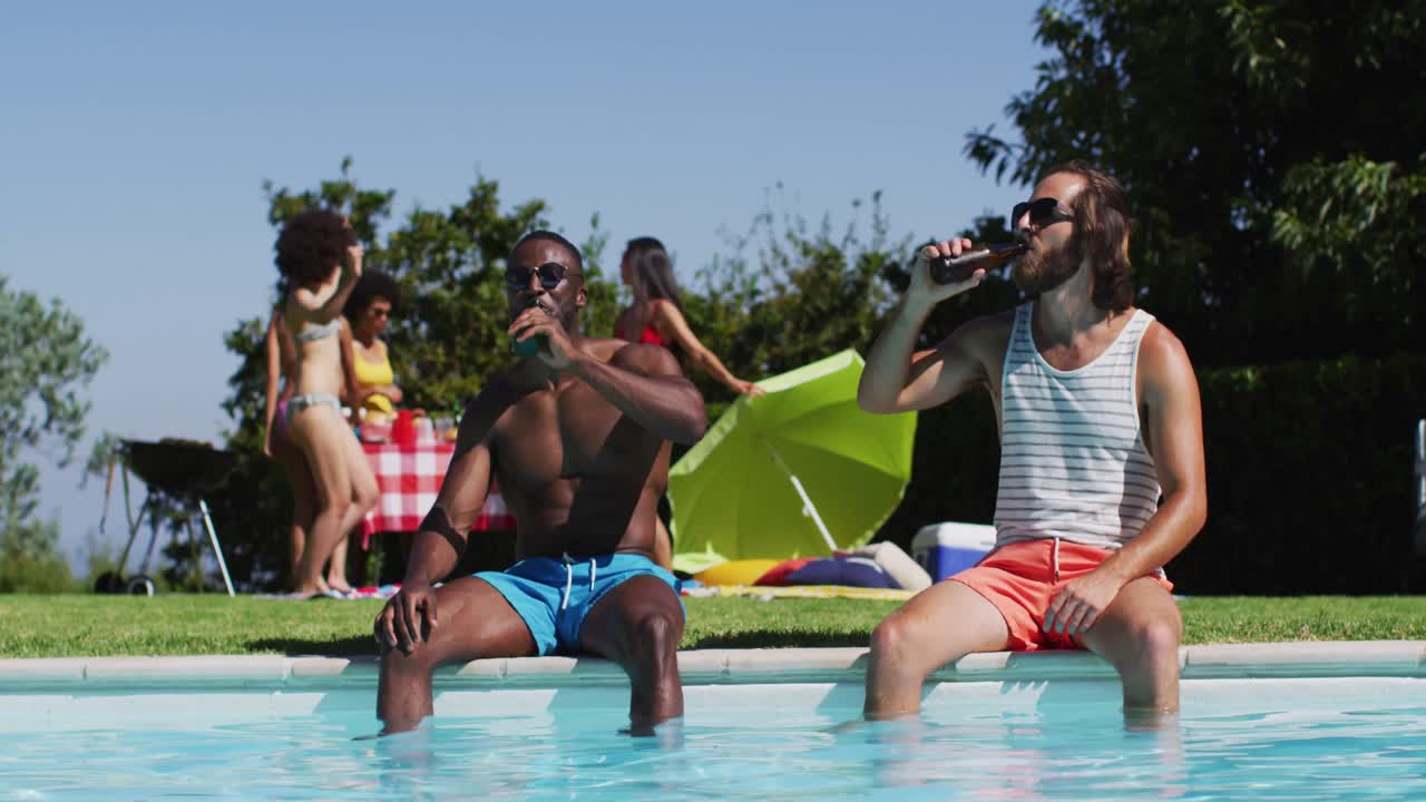 Due amici maschi di diverse origini che brindano e bevono birra seduti a bordo piscina.