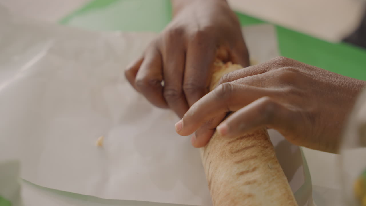 Schwarze Hände rollen einen Tortilla-Wrap während eines Picknick-Dates im Freien. Nahaufnahme des Partners, der einen warmen Shawarma-Wrap auf Papier mit einem grünen Schneidebrett zubereitet. Herzhaftes Hähnchen und frischer Salat fallen heraus. Intime Atmosphäre.