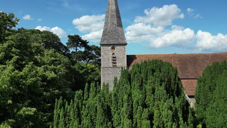 An-aerial-push-in-shot-of-St-John-the-Evangelist-church-in-Ickham,-pushing-in-to-reveal-the-Church-steeple-emerging-from-trees