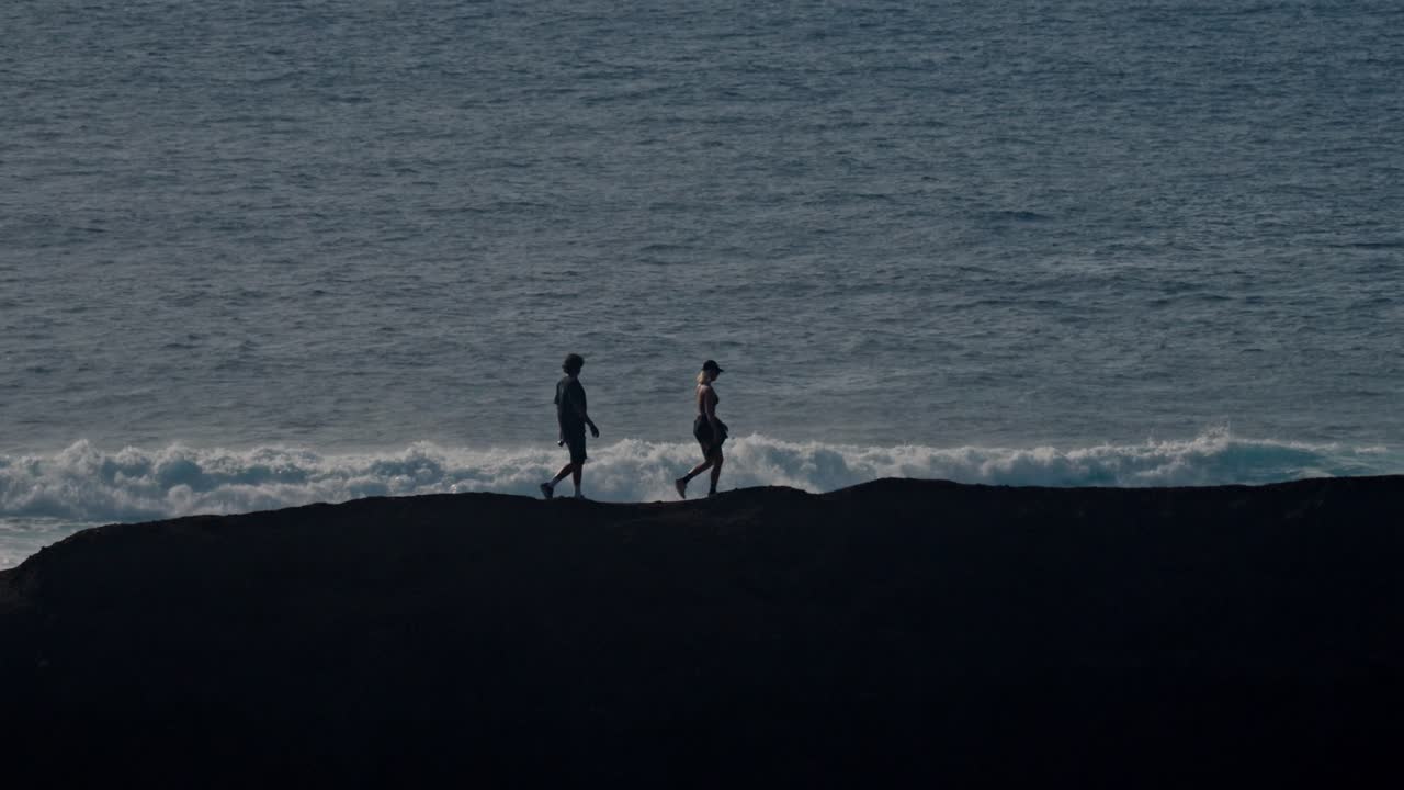 A young couple walks hand in hand along a rugged volcanic cliff overlooking the Atlantic Ocean in Lanzarote, Canary Islands, Spain.