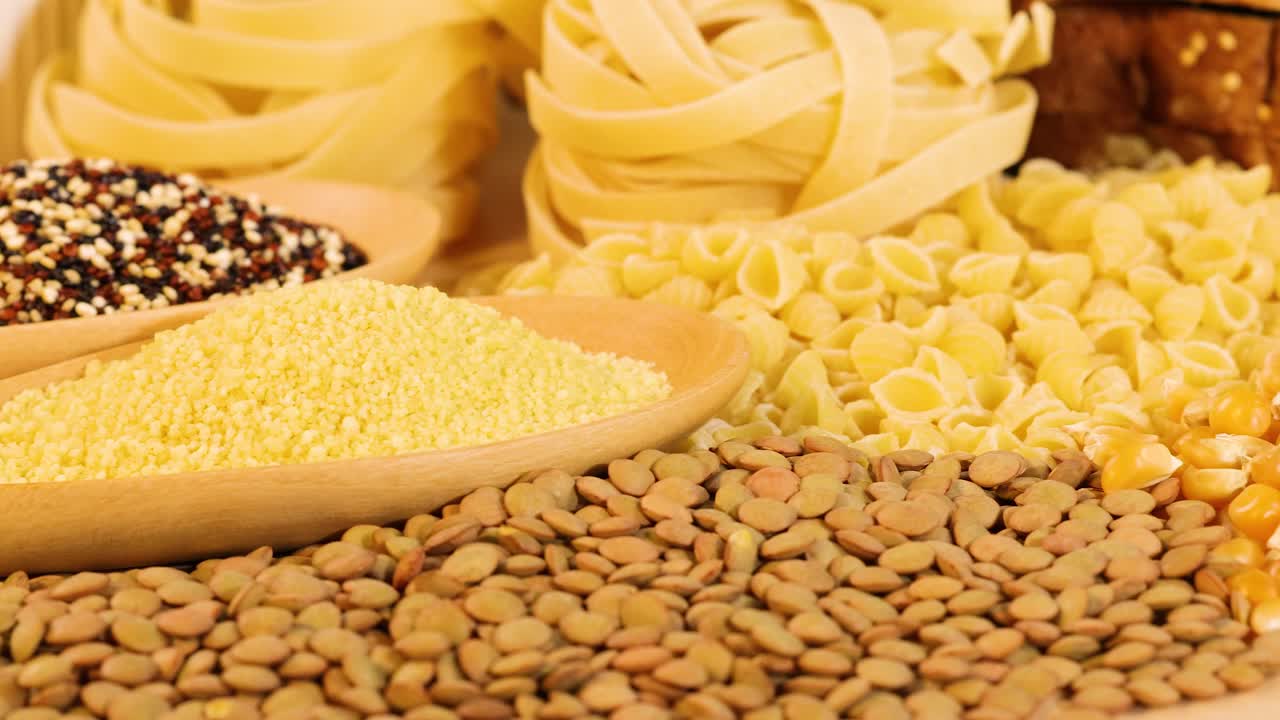 Various grains and pasta displayed with warm lighting, highlighting textures and colors. Includes quinoa, lentils, tagliatelle, and corn