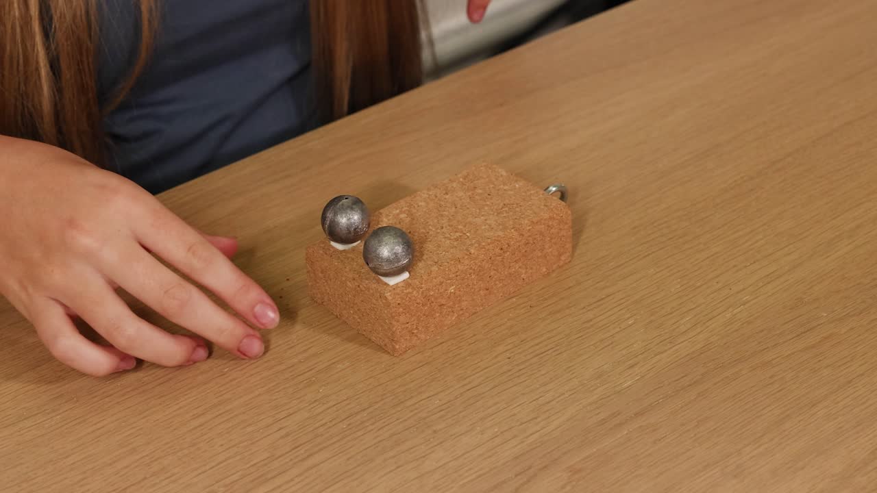 Hands manipulate a cork block with metal spheres on a wooden table under soft lighting, exploring balance and movement
