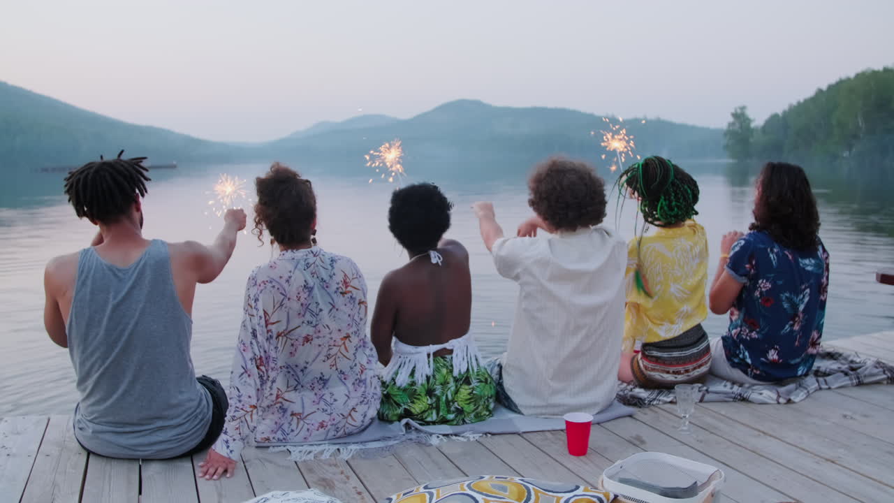 Friends Sitting on Pier and Waving Sparklers at Lake Party