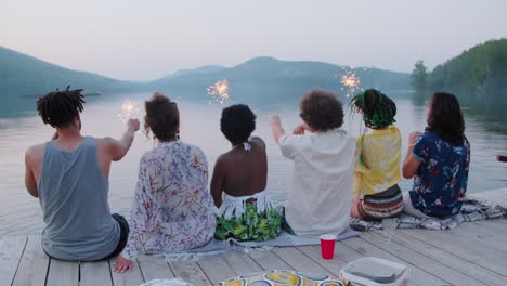 friends sitting on pier and waving sparklers at lake party