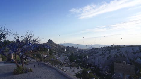 Slow-Motion-Wide-Shot-of-Flock-of-pidgeon-Birds-Flying-Off-Cliff-With-Mountains-and-Air-Balloons-in-Background-in-Uchisar,-Cappadocia,-Turkey