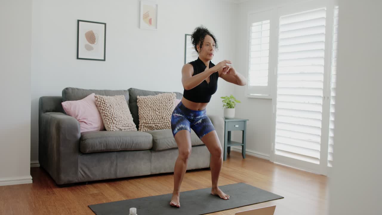 Mulher afro-americana treinando com laptop, fazendo agachamentos no tapete em casa, de roupa esportiva, com garrafa por perto.