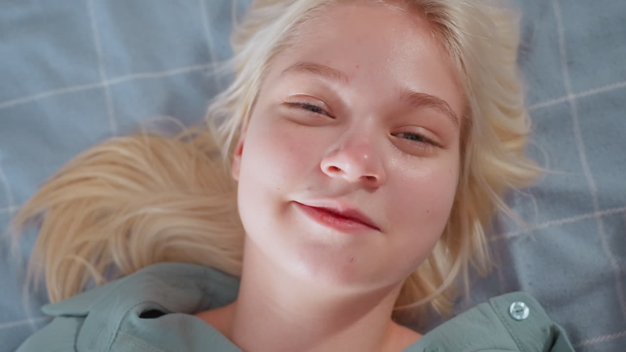 Close up of young blonde woman smiling gently while lying on bed with head turned and eyes open, wearing teal shirt and resting on blue checkered blanket