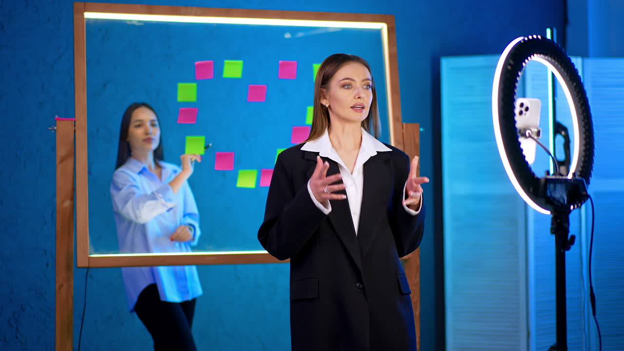 Attractive lady in black jacket talks to camera with ring light. Girl at backdrop puts stickers on the glass wall. Blogging concept.