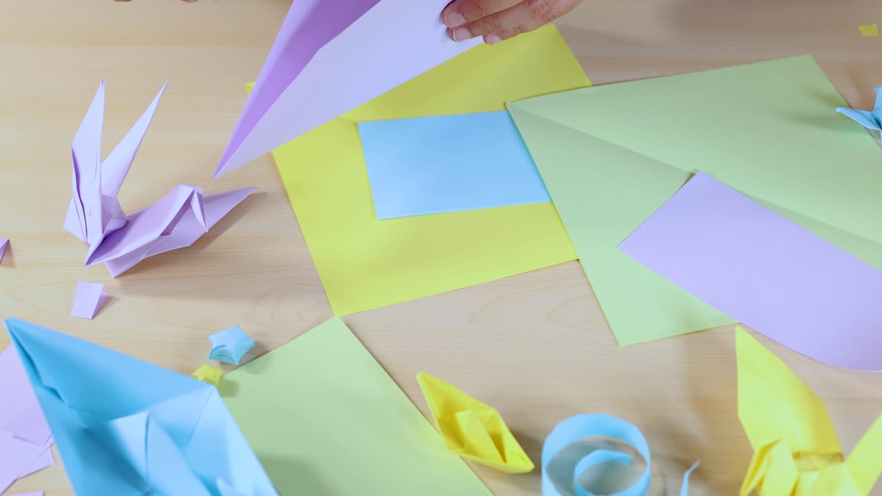 Hands cut pastel paper with a craft knife on a desk surrounded by origami figures