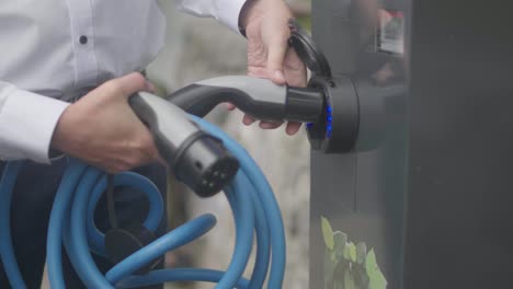 primer plano de un cargador de coche eléctrico moderno conectado a una estación de carga plateada por un hombre que lleva una camisa blanca