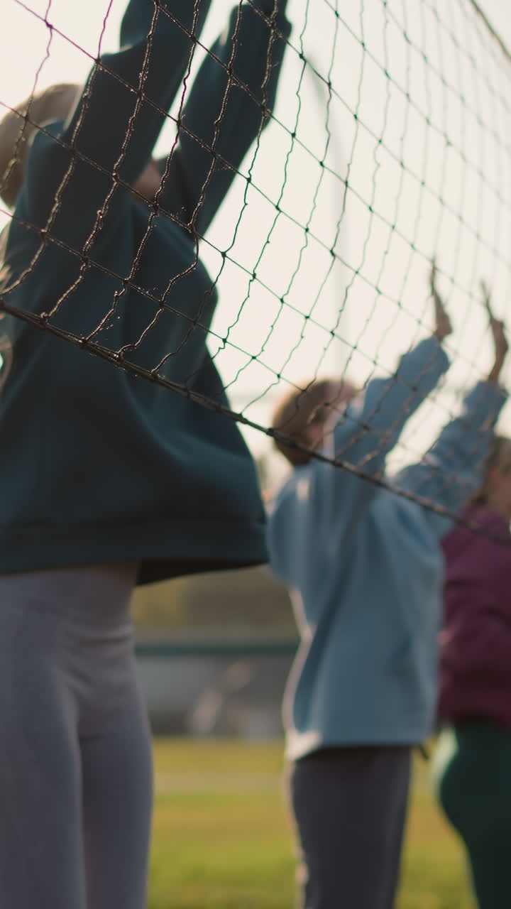 후디를 입은 여성들이 야외에서 볼리볼 훈련에 참여합니다. 코트 근처의 네트, 빛은 금빛 빛을 만니다. 그들은 손을 들고 점프합니다. 배경 특징, 초록색, 그리고 건물.