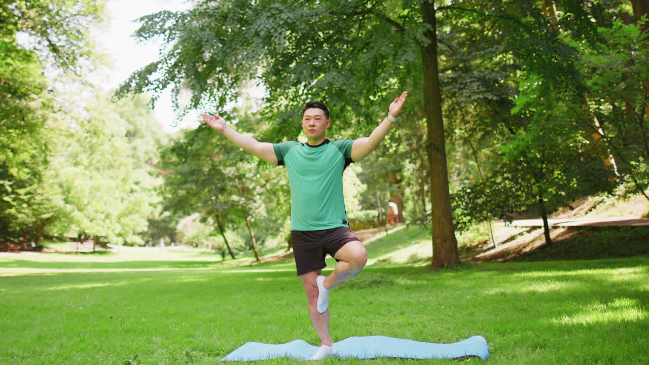 Calm muscular asian japanese sportsman guy balances yogi training on one leg on mat in summer park
