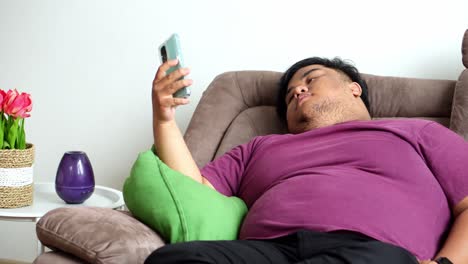 overweight man rests on sofa yawning and covering his mouth while holding smartphone