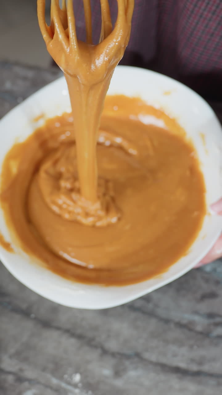 Top-down view of cook holding whisk with thick caramel-colored pap stretching as it drips back into white bowl, flour-dusted countertop and sifter nearby