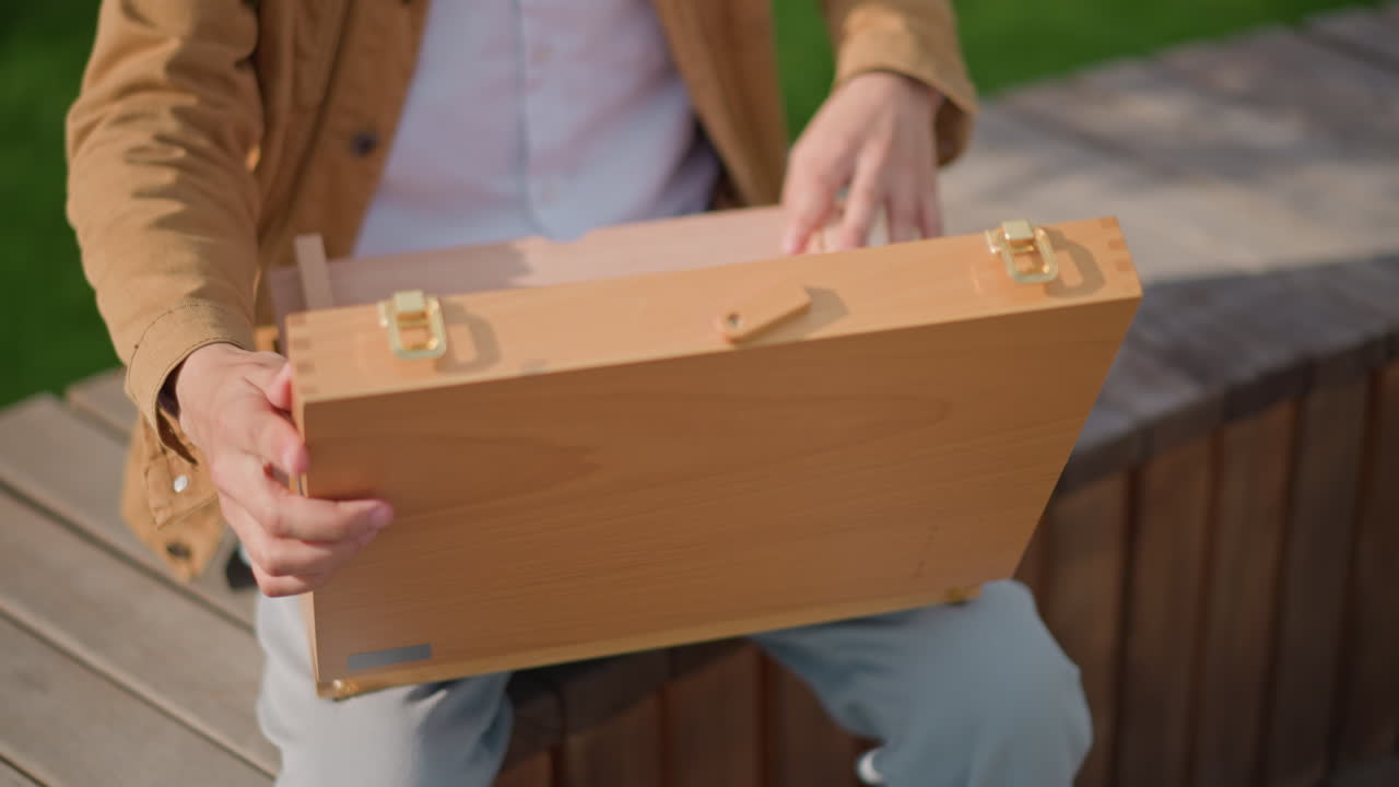 hombre abriendo una caja de dibujo de madera en un parque urbano, preparando un equipo de pintura portátil, interior vacío con compartimentos con bisagras, chaqueta y vaqueros informales, la luz cálida del sol resalta la textura natural de la madera, manos