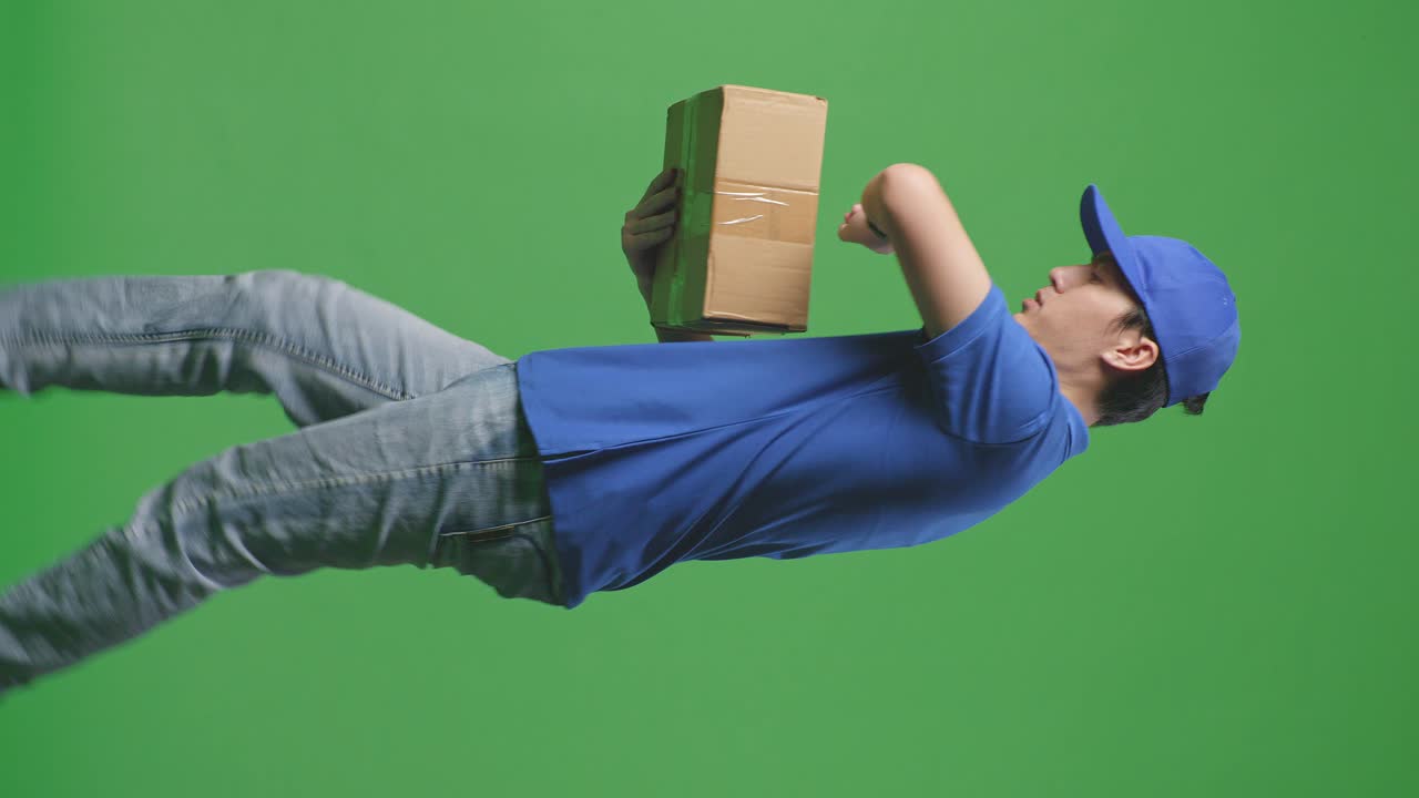 Side View Of Asian Male Courier In Blue Uniform Checking Watch And Running While Delivering A Carton On Green Screen Background In The Studio