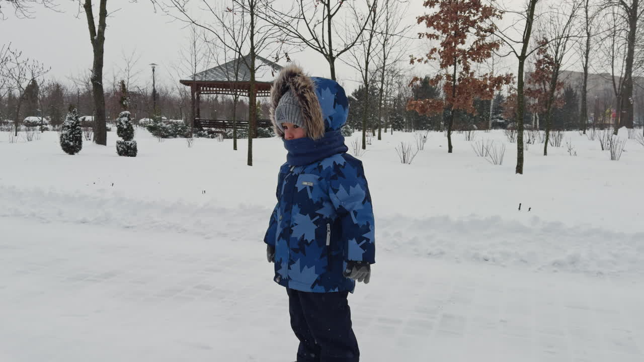 Lovely toddler boy walks by the park on snowy weather. Kid stops, looks around and continues to walk.
