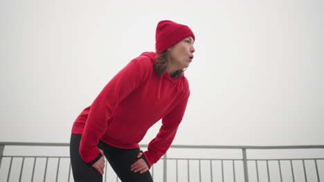 mujer con capucha roja, gorra y leggings negros se inclina, mano sobre pierna, exhalando lentamente con humo visible, cansada, durante el invierno con barandilla de hierro en el fondo