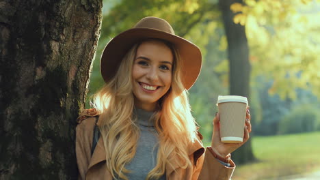 close-up view of blonde woman wearing hat, leaning on a tree, drinking a coffee to go, and looking at the camera with a smile on her face in the park in autumn