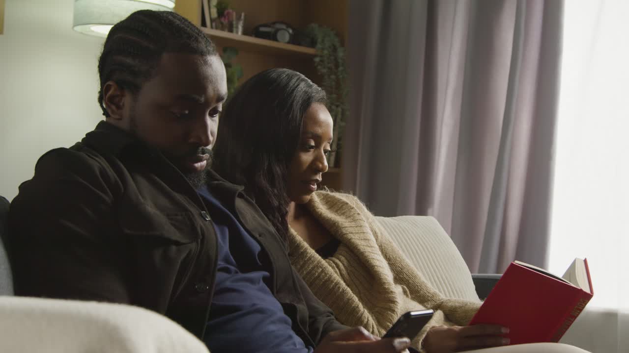 pareja en casa sentada en el sofá en la sala de estar con una mujer leyendo un libro y un hombre mirando el teléfono móvil