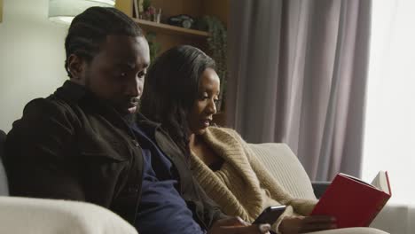 Couple-At-Home-Sitting-On-Sofa-In-Lounge-With-Woman-Reading-Book-And-Man-Looking-At-Mobile-Phone