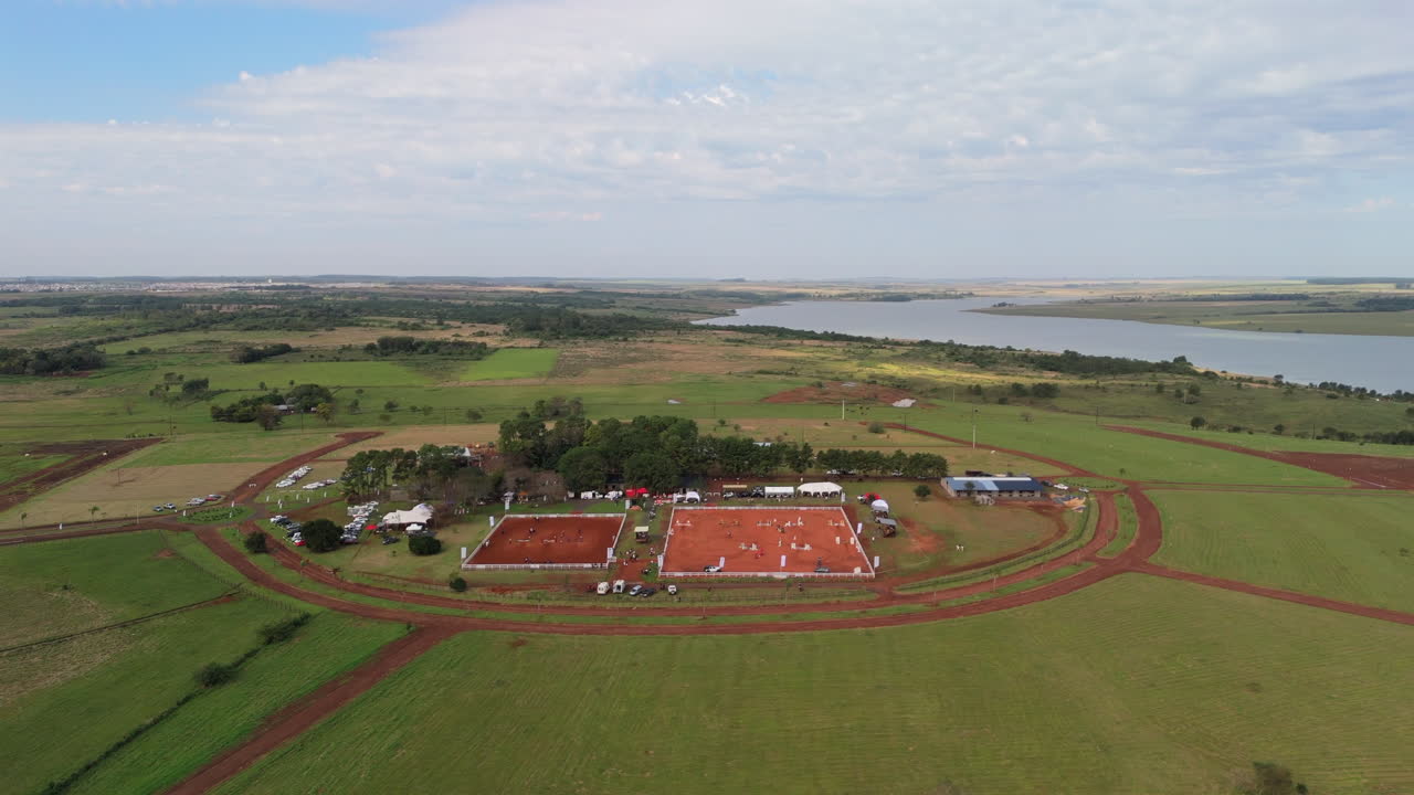 Lateral drone panning over an equestrian club with red clay arenas, surrounded by lush countryside and a distant river in South America.