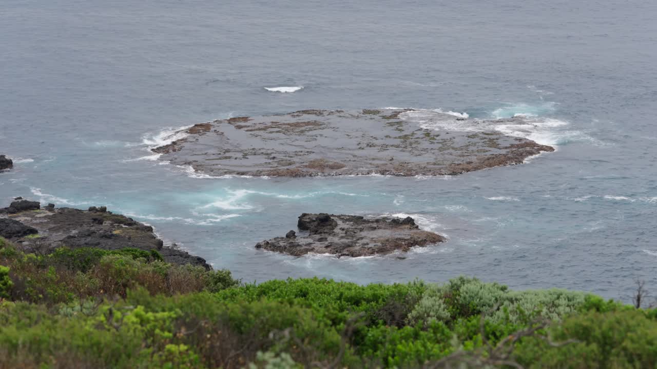 Waves crash powerfully over a rocky island on the shoreline, creating a dramatic and dynamic seascape with water splashing against jagged rocks.