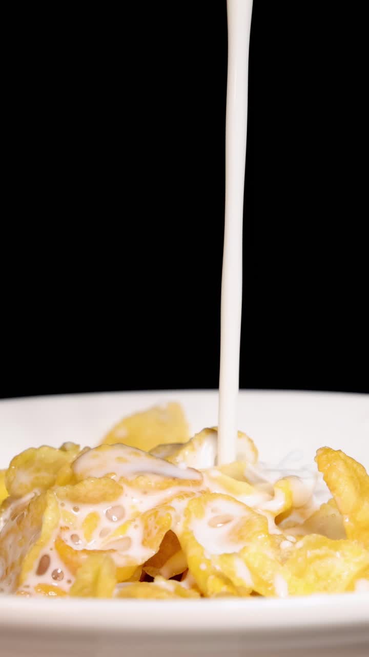 Il latte si riversa a cascata sui cornflakes in una ciotola, su uno sfondo nero, evidenziando il movimento fluido e la consistenza.
