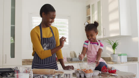 Happy-african-american-mother-and-daughter-wearing-aprons-having-fun-while-cooking-in-kitchen