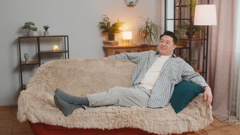 happy carefree middleaged chinese man sitting comfortably on sofa thinking in living room at home