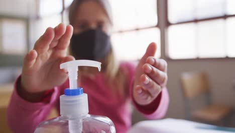 niña con máscara facial desinfectando sus manos mientras está sentada en su escritorio en la escuela