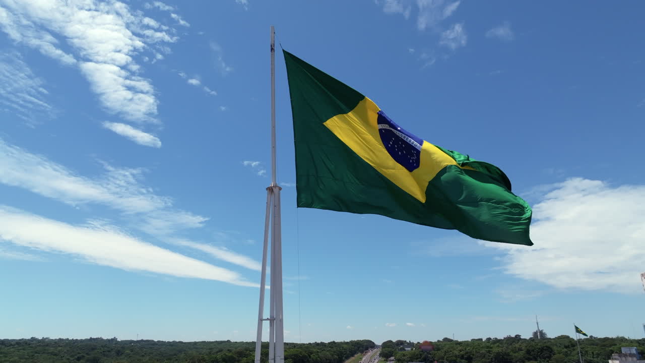 Bandiera brasiliana che sventola su un alto pennone contro il cielo blu e le nuvole in una ripresa aerea con drone. Acquisito in D-Log con colore a 10 bit utilizzando un sensore Hasselblad
