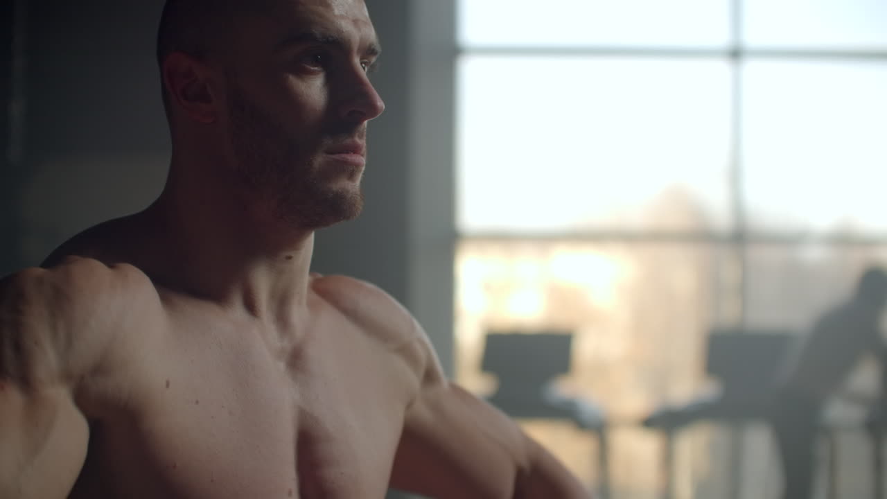 A man without a t-shirt lifts dumbbells in front of the window doing exercises for the shoulder muscles bodybuilder training. Healthy lifestyle and exercises to gain muscle mass. Strength training of a strong willed athlete.