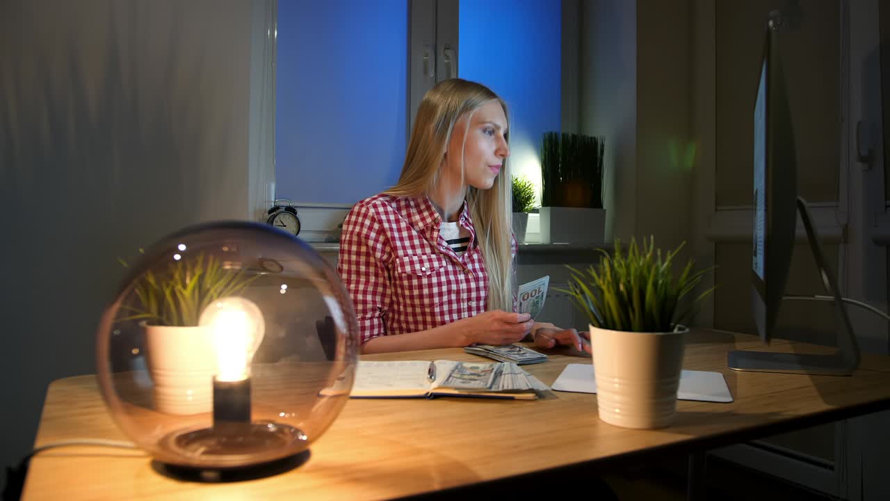 mujer contando dinero en efectivo en el escritorio de trabajo. atractiva mujer rubia en camisa a cuadros sentada por la noche en una habitación con mesa de madera con lámpara y computadora en ella y contando paquete de billetes en las manos.