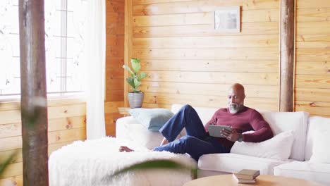 happy senior african american man spending time in log cabin, sitting on sofa and using tablet