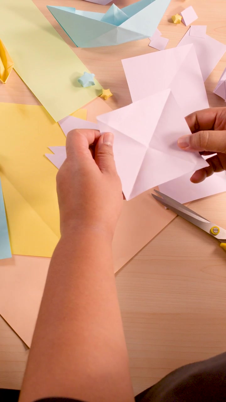 Mani adulte piegano carta bianca per origami su una scrivania color pastello, illuminazione soffusa, vista dall'alto.