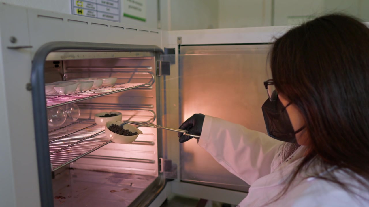 Una scienziata con occhiali e mascherina usa delle pinzette per rimuovere con cura un contenitore in porcellana contenente campioni di terreno da un'autoclave da laboratorio, parte di una ricerca agricola