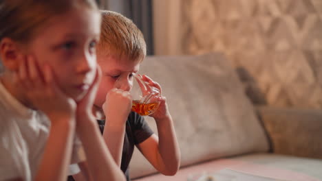 little boy carefully drinks fruit juice from transparent glass cup while girl child sits beside him with hands on cheeks looking paused and thoughtful in cozy indoor setting
