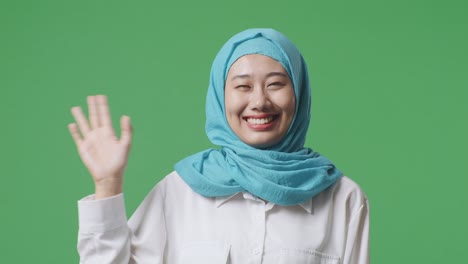 close up of asian muslim woman smiling and waving hand greeting the camera while standing in the green screen background studio