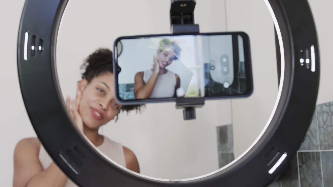 African American woman recording after red dot on phone in bathroom ring light demoing skincare
