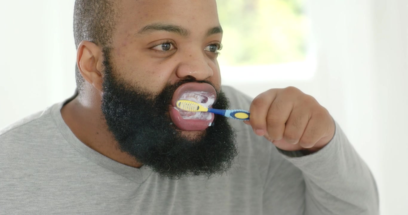 African American man starting with retractor, pressing toothbrush with foam, brushing teeth at sink