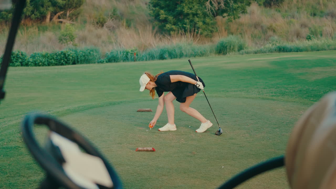 Female golfer bends down to tee up ball on ladies tees, captured from the golf cart driver seat for a unique point of view. Highlights focused preparation with a fresh perspective on the course.