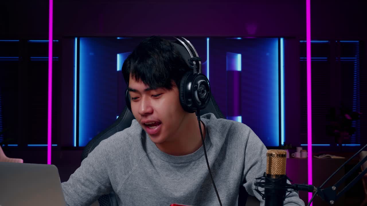 미소 짓는 아시아인 남자 게이머가 헤드폰을 들고 컴퓨터에서 코멘트를 읽으며 레트로 네온 조명으로 조명된 방에서 게임을 하고 있다.