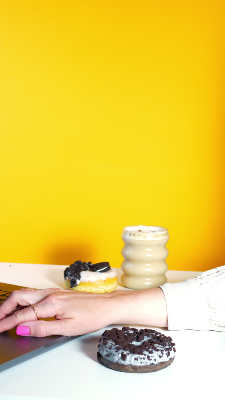A hand with pink nails types on a laptop and reaches for a chocolate cookie crumble donut. iced coffee, another donut on a clean white desk with a vibrant yellow backdrop blending work and indulgence.