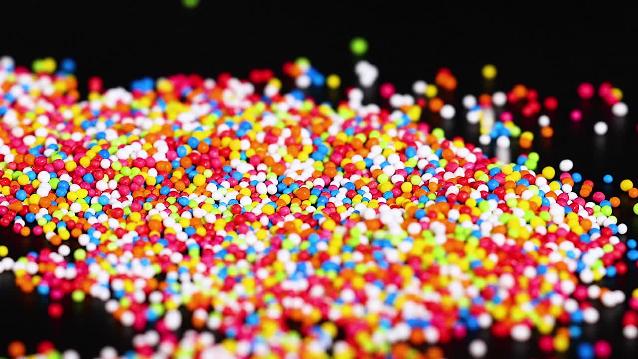 A hand interacts with colorful sprinkles scattered across a dark background, creating dynamic patterns and textures.