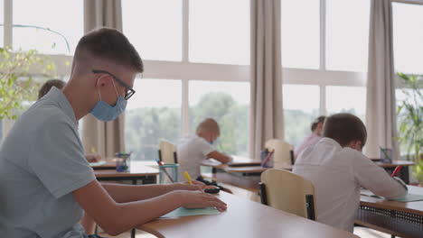grupo multirracial de niños con máscaras faciales trabajando en clase escribiendo y escuchando las explicaciones del maestro en el aula