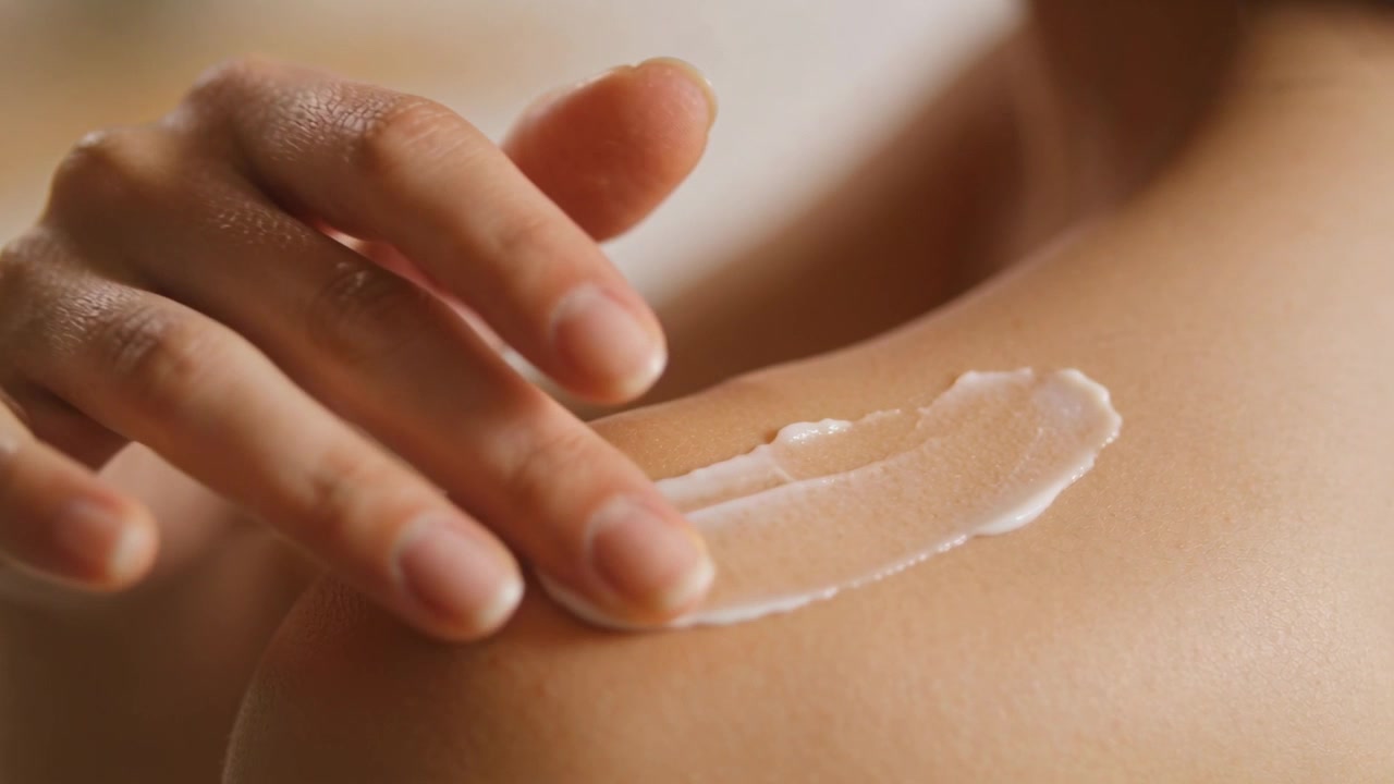 Spreading woman's hand pressing dollop, smoothing white cream on shoulder for hydration at home