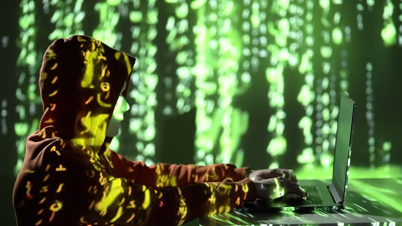 Masked hacker in hoodie types on laptop in dark room, lit by green digital code. 4K slow-motion shot symbolizing cybercrime, data breaches, and online anonymity in a tech-driven world.