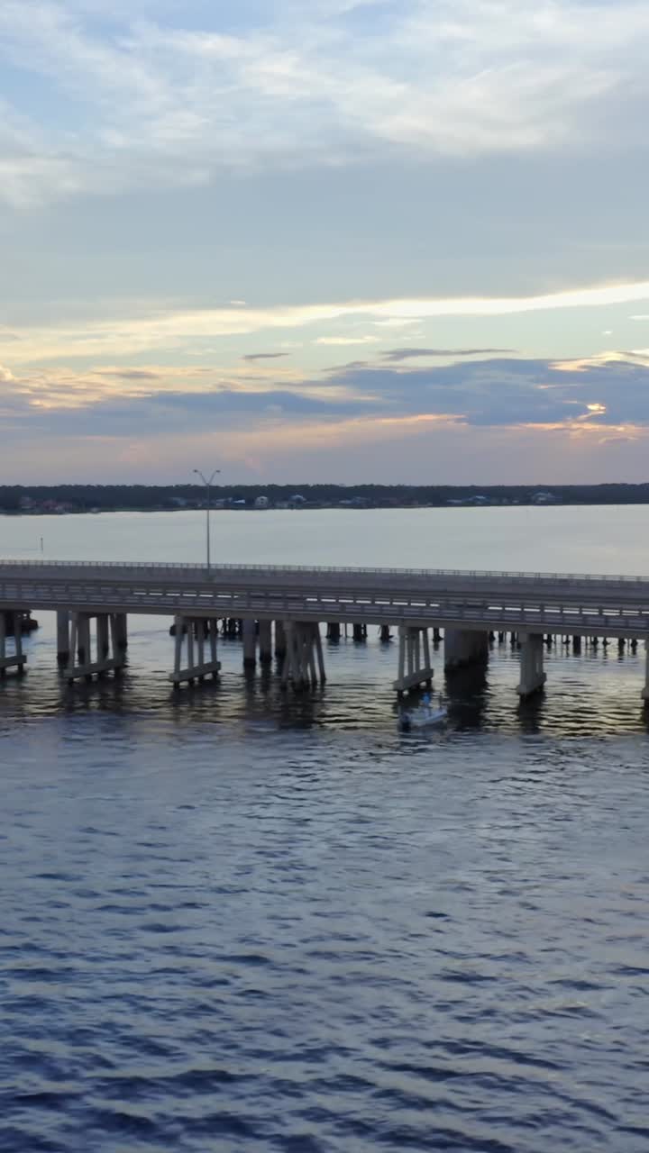 A concrete bridge stretches over still blue bay waters as the warm hues of the evening sky reflect gently on the surface, capturing a peaceful coastal moment