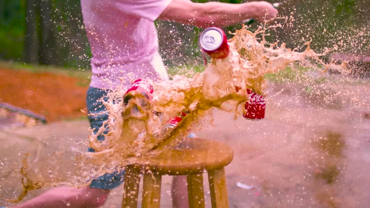 Three red Coca-Cola soda cans being sliced in half by a machete or a small sword in slow motion. The cans are on a small table or a wooden chair and filmed during summer time with trees in the back.